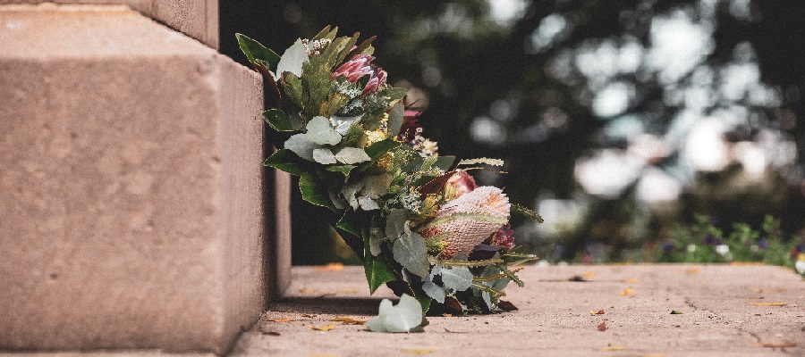 Ein vakker krans med ulike blomar og grøne blad, plassert ved ein steinmur. Dei jordnære tonane og detaljane i blomane gir eit naturleg og verdig uttrykk, med ein uskarp bakgrunn som skapar djupne i biletet. Foto.